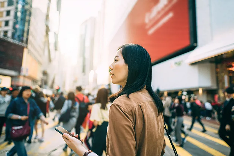 Young woman checking on mobile phone while crossing street and commuting in busy downtown city street