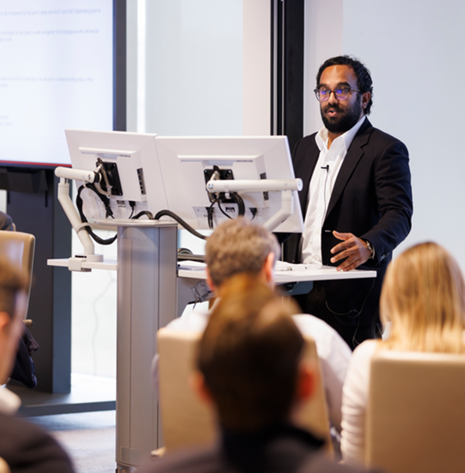 Presenter with glasses and dark suit standing at a lectern, engaging a seated audience.