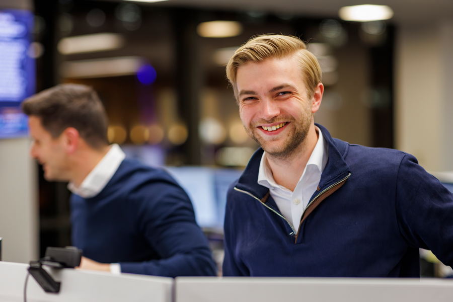 Smiling young man with blonde hair and beard in a navy sweater, with colleagues and monitors blurred behind.