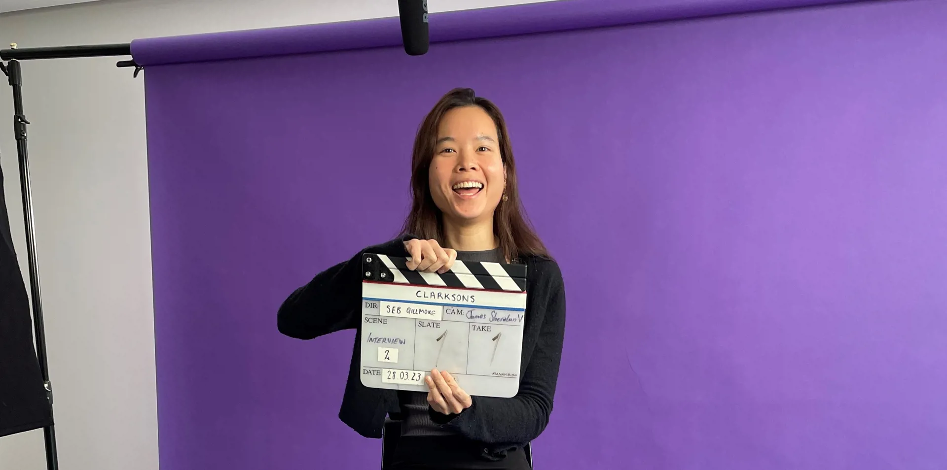 woman sitting on a stool behind a purple background, smiling, holding a clapper