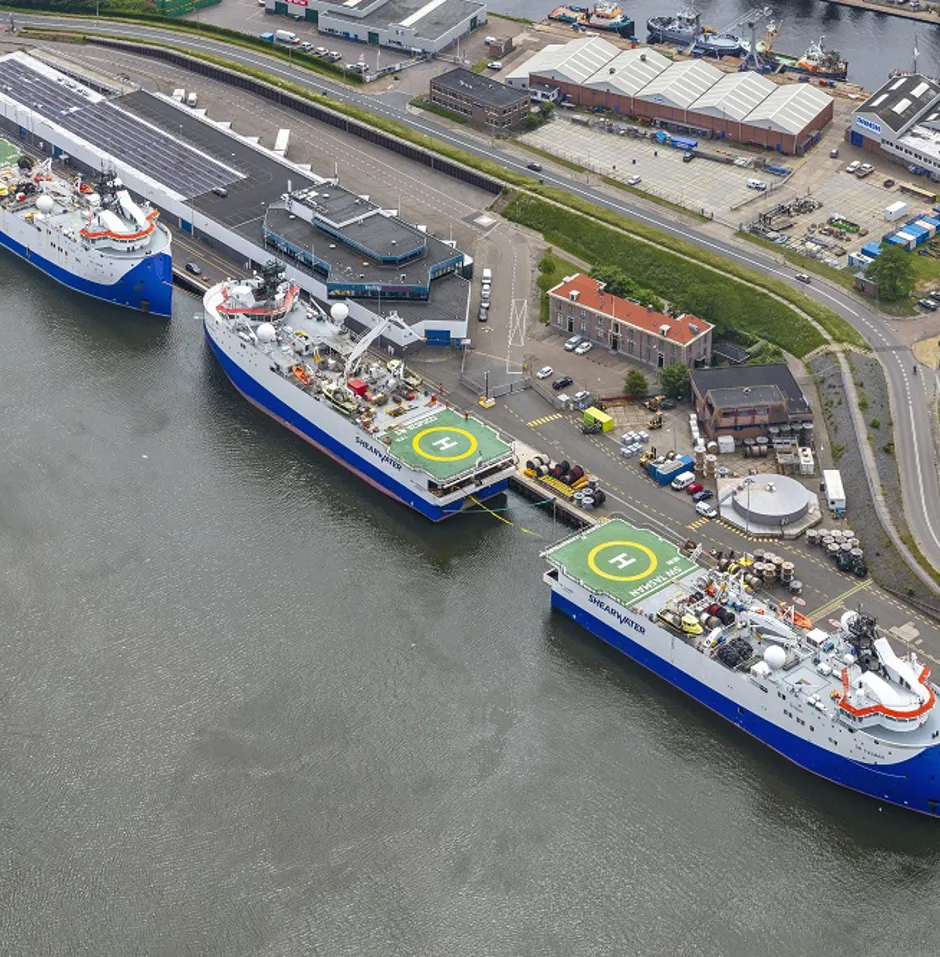 Three ships docked at a harbor with warehouses.