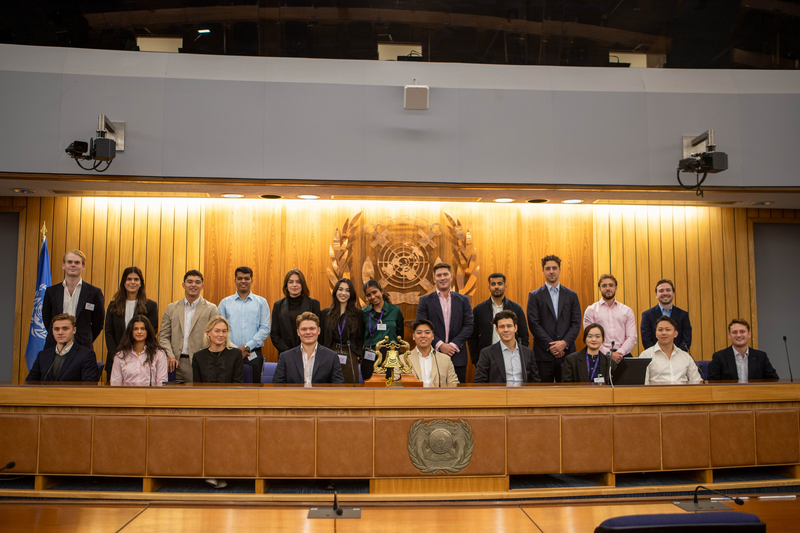 Group in business attire posing in a formal room with a United Nations emblem backdrop.