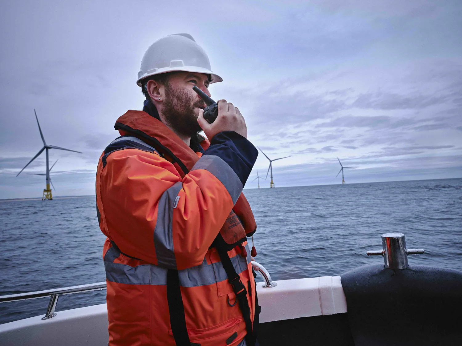 Man in high vis jacket and hard hat standing on board a vessel with windfarms on the horizons. 