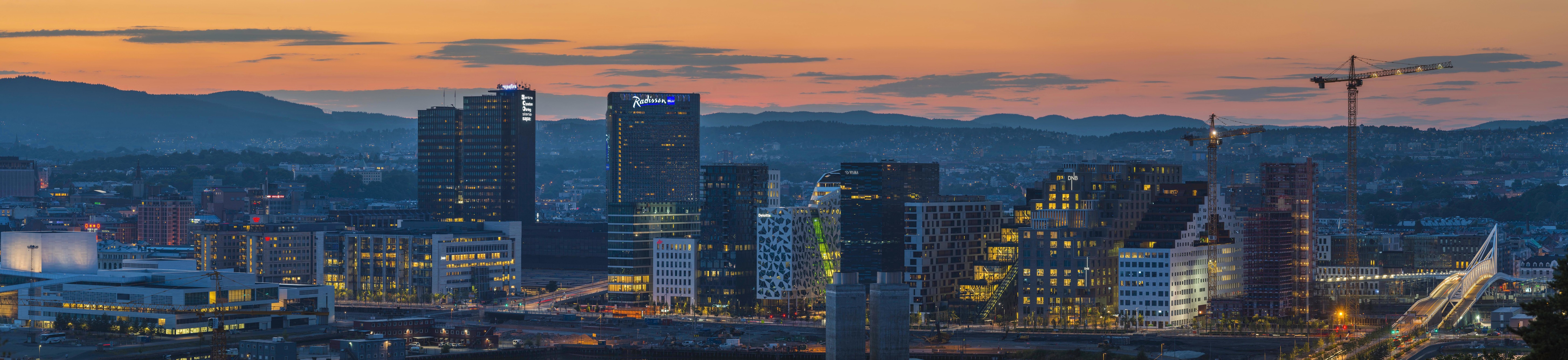 Panoramic view of modern city skyline at dusk with illuminated high-rise buildings and distant mountains.