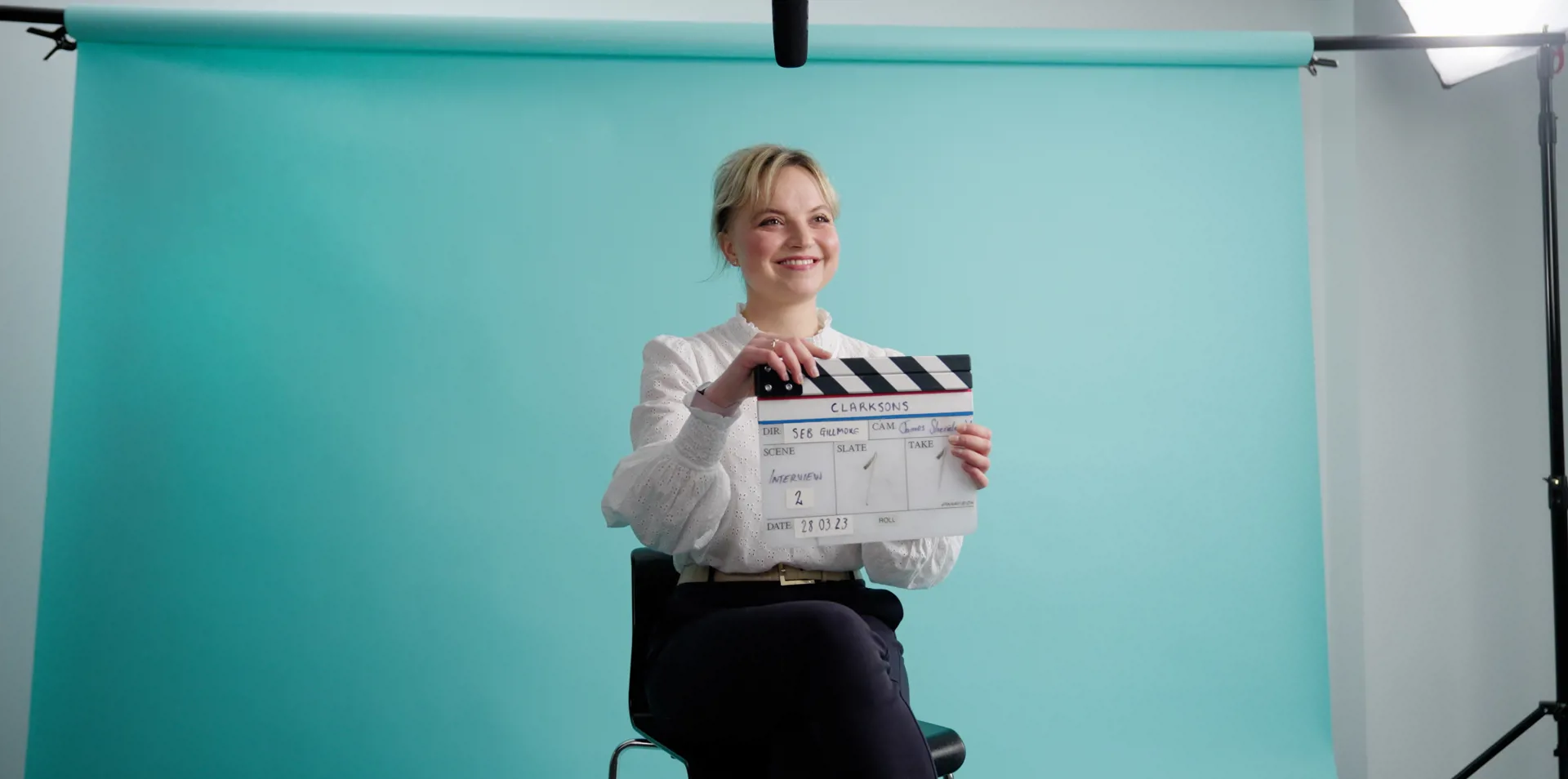 woman sitting on a stool behind a blue background, holding a clapper