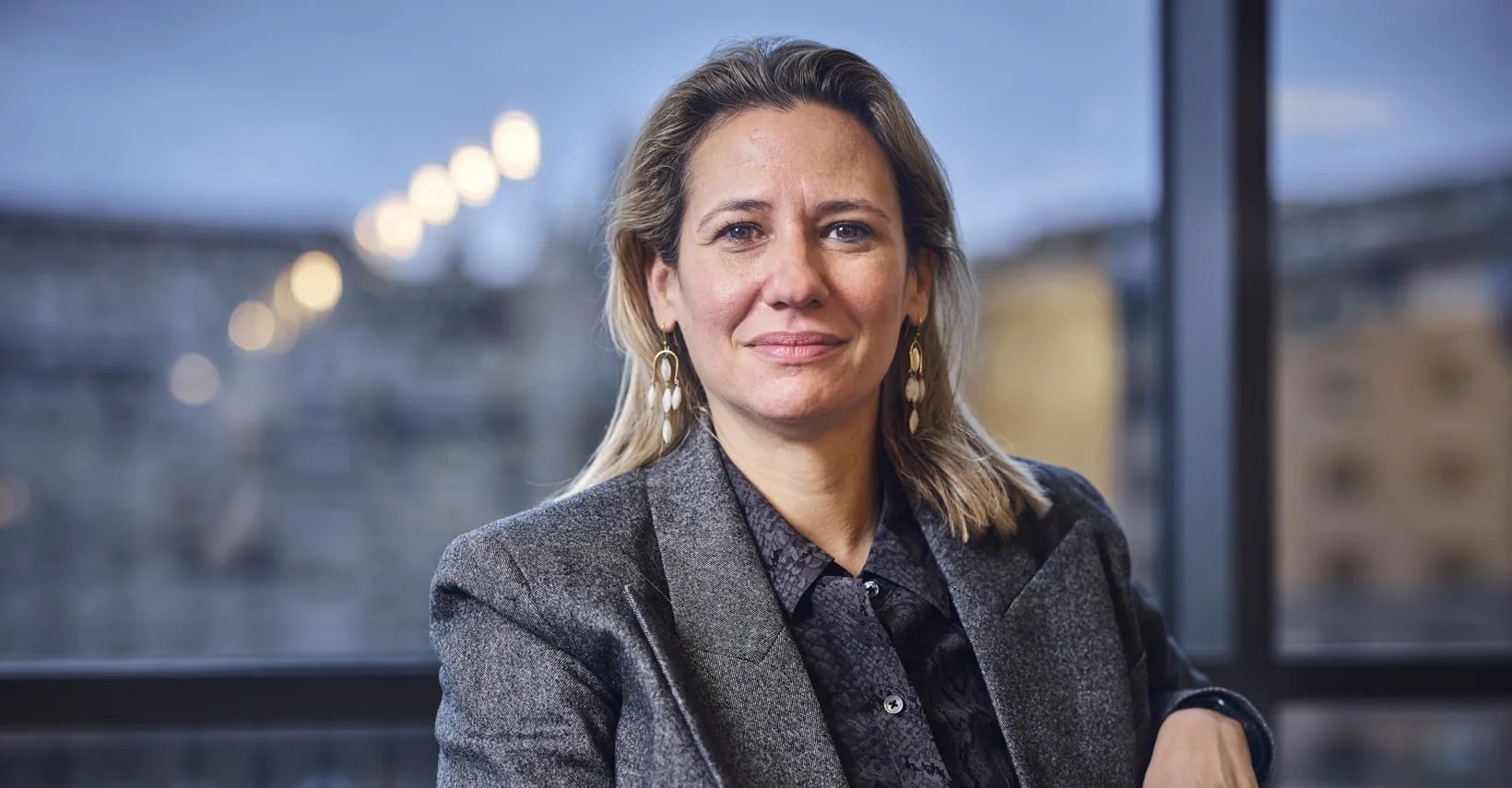 blonde woman smiling at camera, sitting on chair with arm on side