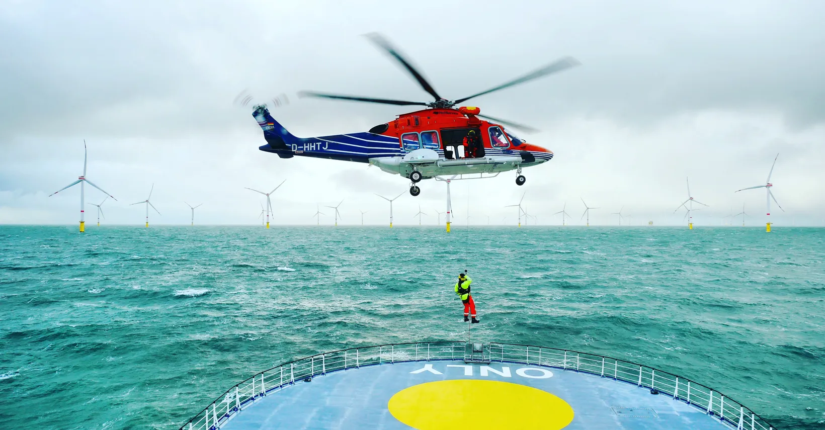 Helicopter hovering above a ship near wind turbines.